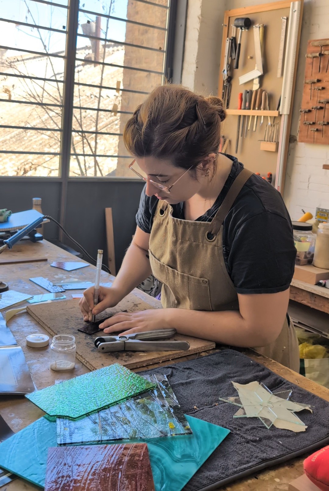 Luna cortando una pieza de vidrio sobre la mesa de trabajo, rodeada de vidrio de colores